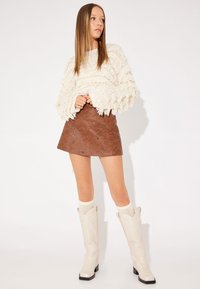 Maglione bianco con texture e dettagli frangiati, gonna in finta pelle marrone con dettagli a occhiello, stivali crema fino al ginocchio e calze leggere in maglia.