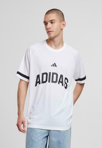 Bela atletska majica s črnim tiskom "ADIDAS", raglan rokavi in črne poudarjene črte na rokavih. Mehka, zračna tkanina.