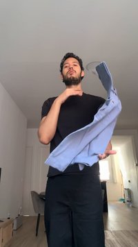 Man with beard and curly hair stands indoors, wearing black clothes, holding and shaking a light blue shirt in a room with furniture and a window.