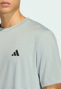 Svetlosivé krátke tričko s krátkym rukávom z mäkkej látky, s čiernym logom s tromi pruhmi na ľavej strane hrudníka. Okrúhly výstrih a voľný strih.