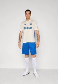 Camisola creme com detalhes azuis, apresentando uma gola polo, logótipo "Betano" e emblema do clube. Combinada com calções azuis e meias brancas até ao joelho.