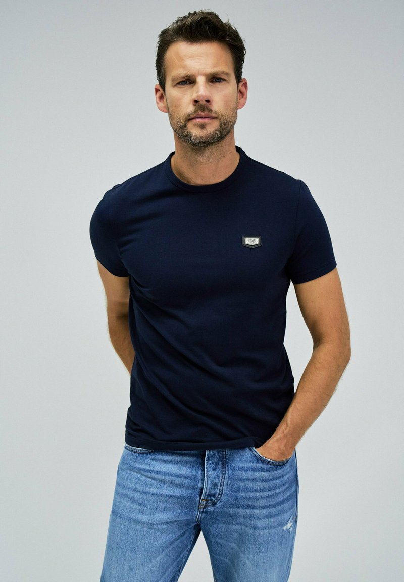 Camiseta de manga corta azul marino hecha de algodón, con un pequeño parche de logo en el pecho. Textura suave, cuello redondo y diseño ajustado.