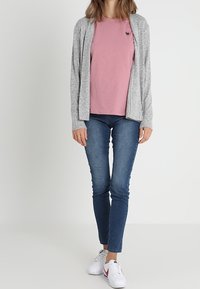 Cardigan grigio aperto sopra una t-shirt rosa, abbinate a jeans blu aderenti e sneaker bianche. Caratteristiche notevoli includono un piccolo logo sulla maglietta.