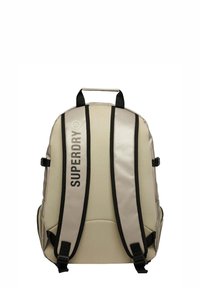 Zaino beige con cinghie e dettagli neri, con il logo "SUPERDRY" in caratteri audaci, progettato per durabilità e funzionalità.