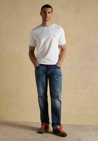 Jeune homme debout les mains dans les poches, portant un t-shirt blanc, un jean bleu et des chaussures marron avec des lacets orange vif, sur un fond beige.