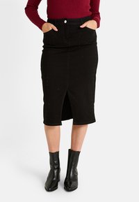 Falda lápiz de denim negro con abertura lateral, adornada con acentos de strass, dos bolsillos frontales y un diseño ajustado. Combinada con botas negras de tobillo.