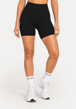 Femme portant un short de vélo taille haute à imprimé léopard noir, des chaussettes blanches aux chevilles et des chaussures de sport blanches New Balance, debout sur un fond blanc.