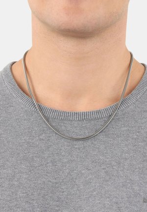Silberne Schlangenkette, die auf einem grauen Pullover liegt. Die Halskette hat eine glatte, reflektierende Oberfläche und ein einfaches, rundes Design.