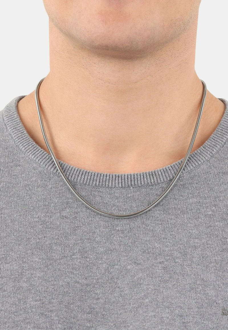 Silberne Schlangenkette, die auf einem grauen Pullover liegt. Die Halskette hat eine glatte, reflektierende Oberfläche und ein einfaches, rundes Design.