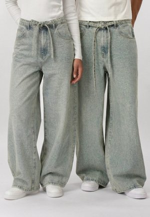 Két ember világosra mosott, széles szárú farmernadrágot visel, melynek dereka megkötős, fehér tornacipőben, egymás mellett állnak.