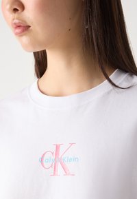 Balta medvilninė marškinėlė su apvaliu iškirpimu. Priekyje yra rožinės spalvos "CK" ir mėlynos spalvos "Calvin Klein" prekės ženklai, užrašyti paprastu ir šiuolaikišku šriftu.