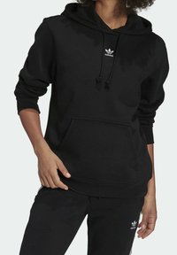 Svart hoodie med känguruficka, dragsnöre vid huvan och liten vit Adidas-logga på bröstet. Mjuk tyg med ribbade muddar.