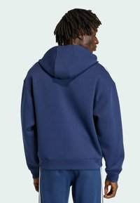 Muž s tmavou pleťou a dredmi, oblečený v námornícky modrej mikine s kapucňou a zodpovedajúcich nohaviciach s bielymi pruhmi, otočený chrbtom k jednoduchému pozadiu.