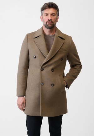Dobbeltbrystet brun uldfrakke med klapkrave, store sorte knapper og sidelommer, båret over en strikket sweater. Sorte bukser synlige.
