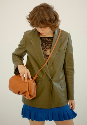 Blazer de cuero verde oliva, diseño de doble botonadura. Vestido de punto azul con dobladillo con volantes. Bolso de cuero marrón cruzado con detalle de cremallera.