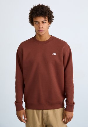 Sudadera marrón con cuello y puños acanalados, que presenta un pequeño logotipo blanco en el pecho izquierdo. Textura suave y corte relajado.