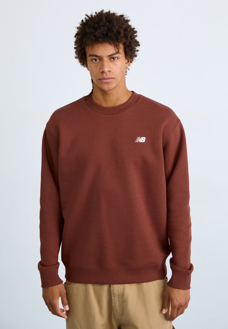 Brauner Pullover mit geripptem Kragen und Bündchen, ausgestattet mit einem kleinen weißen Logo auf der linken Brust. Glatte Textur und entspannter Schnitt.
