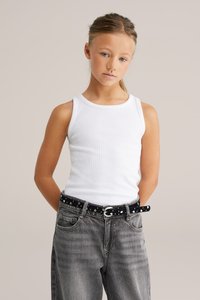 Top de tirantes blanco acanalado combinado con unos vaqueros de mezclilla grises y holgados. Cinturón negro con acentos metálicos y lunares blancos. La modelo tiene el cabello rubio liso.