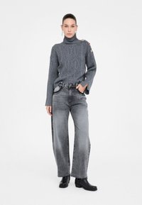 Donna che indossa un maglione a collo alto grigio a trecce, pantaloni jeans grigi sbiaditi a gamba larga con rifiniture nere e stivali neri alla caviglia su uno sfondo bianco.
