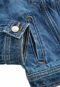 Dettaglio di giacca di denim con tessuto blu e finitura sbiadita, cuciture a contrasto, bottone a pressione argentato e tasca laterale.