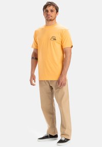 Yellow cotton t-shirt with short sleeves, round neckline, and a small black graphic on the left chest. Worn with khaki pants and black shoes.
