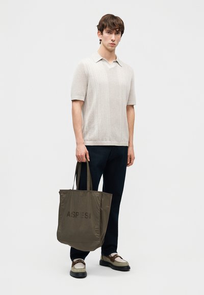 Jeune homme portant un polo en maille beige à manches courtes, pantalon sombre, mocassins bicolores, tenant un sac fourre-tout olive avec le logo "ASPESI".