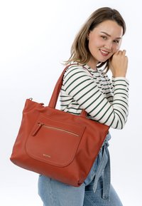 Punase tekstuuriga nahast kandekott ees luku tasku, kahe käepideme ja avara disainiga, mida täiendavad kuldsed metalldetailid.