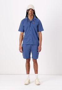 Chemise à boutons bleue et shorts assortis, en tissu doux. Logo notable sur la chemise, avec une coupe décontractée et un design uni.