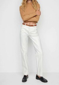 Weiße Jeans mit geradem Bein und glatter Textur, mit Vordertaschen und braunem Ledergürtel. Kombiniert mit einem kurzen, hellbraunen Pullover und schwarzen Schuhen.