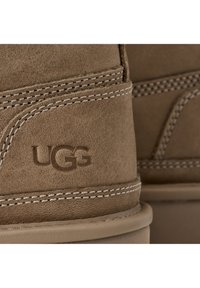 Bota de ante marrón con forma redondeada, que presenta un detalle de costura, suela de goma y el logo de UGG grabado en el lateral.