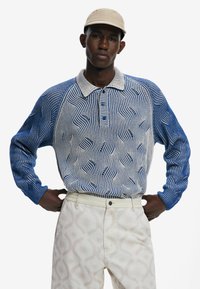 Polo cu dungi albastre și albe, cu mâneci lungi, având un model texturat de valuri, dispune de guler și nasturi, fiind asociat cu pantaloni bej cu un model subtil.