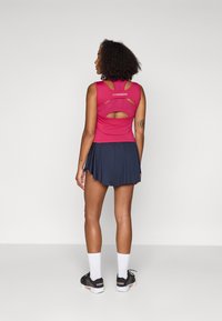 Top sportivo rosa con dettagli a taglio sul retro, abbinato a una gonna plissettata blu navy. Il modello indossa scarpe da allenamento nere e calzini bianchi.
