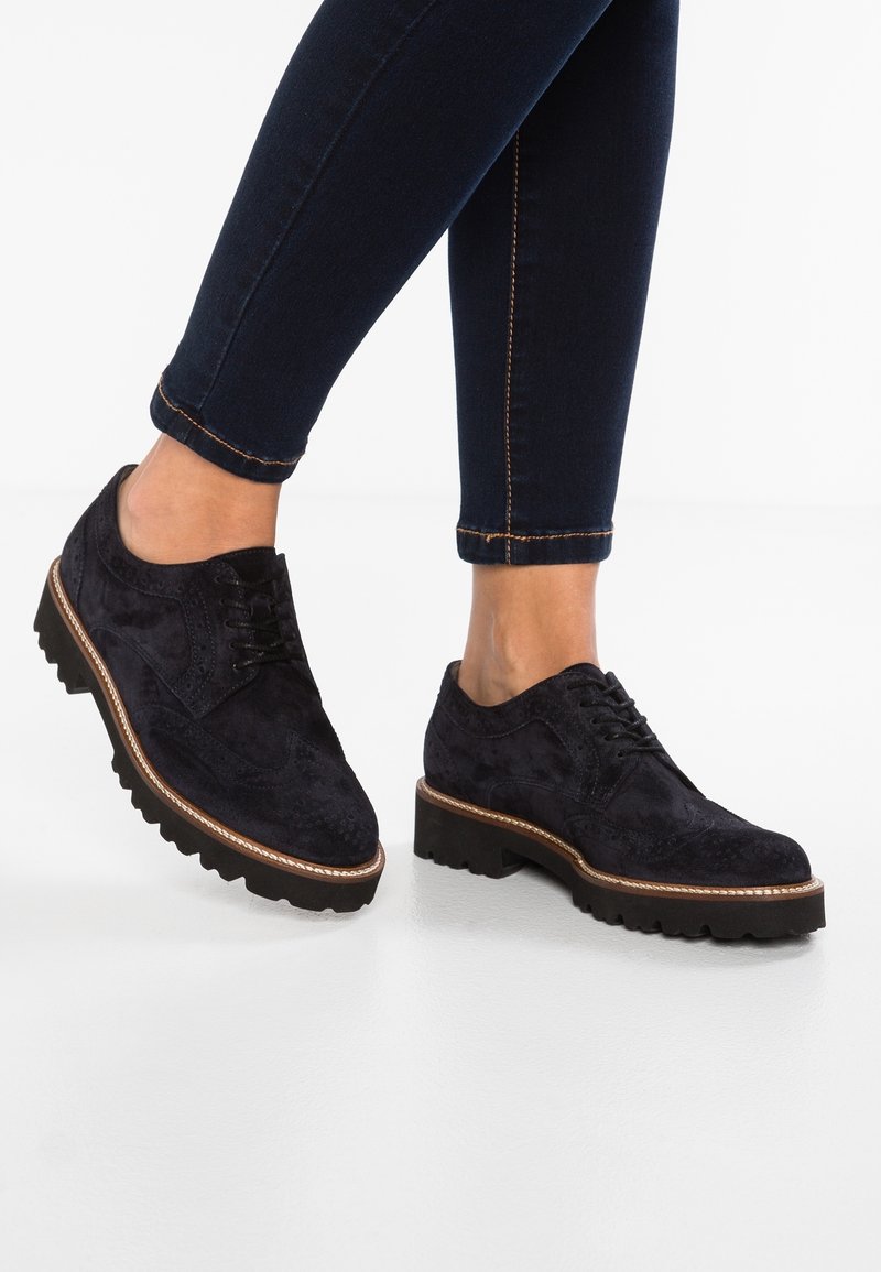 Brogues en daim noir à lacets avec semelles épaisses à crampons portés avec un jean skinny foncé, montrant les jambes du mollet jusqu'aux pieds sur fond blanc.