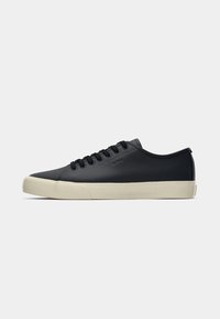 Baskets basses en cuir noir avec lacets noirs, nom de marque embossé et semelle en caoutchouc blanc cassé sur fond blanc.