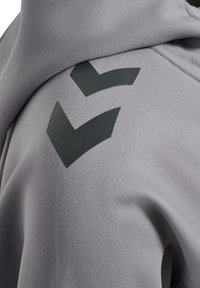 Grå hoodie gjord av mjukt material, med en svart chevrondesign på axeln och en texturerad, slät yta.