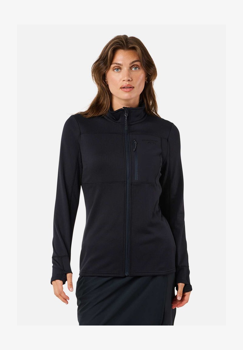 Vrouw met lichte huid en bruin haar, die een zwarte jas met rits en een borstzak draagt, staand tegen een witte achtergrond.