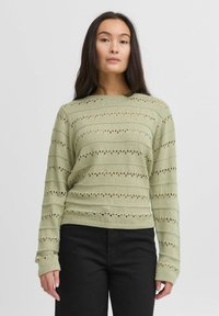 Ljusgrön stickad tröja med horisontella urklippta mönster, långa ärmar och rundad halsringning, tillsammans med svarta byxor.