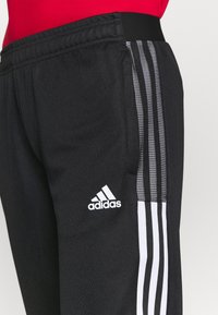 Černé sportovní kalhoty s třemi bílými pruhy po straně, elastickým pasem a malým bílým logem Adidas na dolní části nohy.