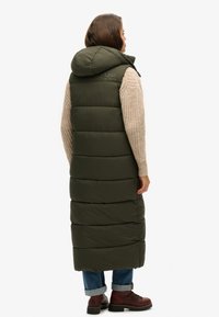 Langer olivgrüner Puffer-Weste mit Kapuze, die über gesteppten Abschnitte und eine strukturierte Oberfläche verfügt, kombiniert mit einem cremefarbenen Strickpullover und Jeans.