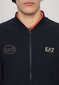 Mornarska dukserica na zip s rebrastom teksturom, narančastim ovratnikom i zlatnim EA7 logotipima na lijevoj strani prsnog dijela i području zatvaranja na prednjoj strani.