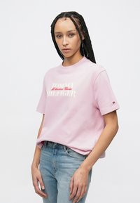 Ανοιχτόροζ βαμβακερό μπλουζάκι με στρογγυλή λαιμόκοψη και κοντά μανίκια, με λευκό και κόκκινο σχέδιο λογότυπου "TOMMY HILFIGER" στο στήθος.