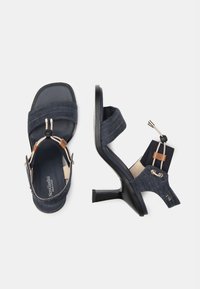 Mørkeblå denim højhælede sandaler med firkantet tå, elastiske stropper og en brun læderdetalje. Har et slankt, minimalistisk design.