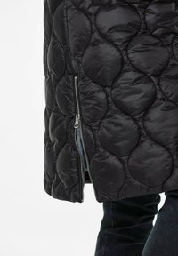 Schwarze gesteppte Jacke mit einem strukturierten Muster aus kreisförmigen Abschnitten und einem silbernen seitlichen Reißverschluss. Das Material wirkt glänzend und glatt.