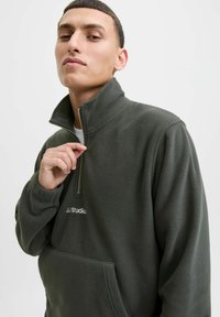 Young man with short hair wearing dark green fleece jacket partially zipped, holding zipper near collar, neutral light background.