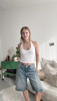 Top de halter branco e calções de ganga azul claro com um acabamento desbotado. Quarto com um armário verde e plantas decorativas ao fundo.