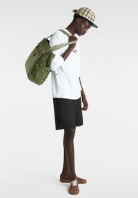 Olivgrüner Rucksack mit mehreren Taschen, über einer Schulter getragen, kombiniert mit einem weißen Langarmshirt, schwarzen Shorts und braunen Schuhen.