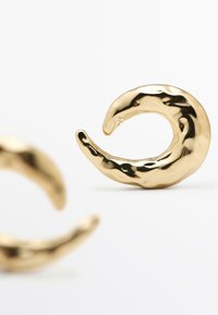 Náušnice z kovu se zlatavým tónem ve tvaru zakřivených vln s texturovaným povrchem, které mají lesklý, odrážející se povrch a hladké okraje.