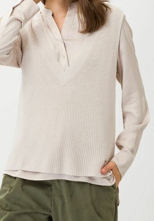 Femme portant un gilet sans manches en maille beige clair à col en V sur une chemise boutonnée beige pâle avec un pantalon vert olive, la main dans la poche.