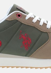Olivno zeleni športni copat z bež akcenti, ki ima rdeč logotip igralca polo. Teksturirano obdelavo, bele vezalke in gumijast podplat dopolnjujejo oblikovanje.