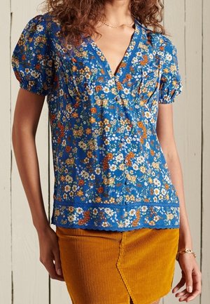 Kvinde iført en blå blomsterprint kortærmet bluse med puffede ærmer og et okker gult kordnøgle-nederdel med en frontslids.